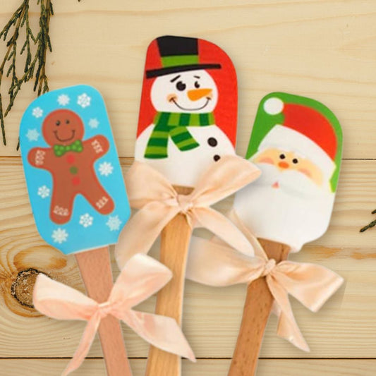 Three silicone spatulas with wooden handles displayed. Gingerbread man, Snowman, and Santa patterns are shown.