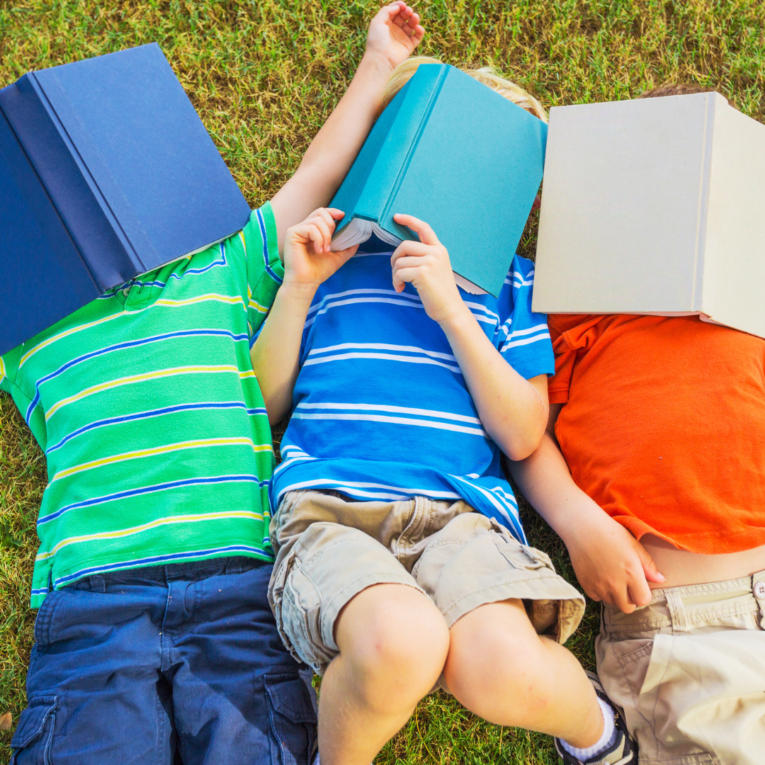 3 grade school boys lay on the grass on a sunny day with books over their faces