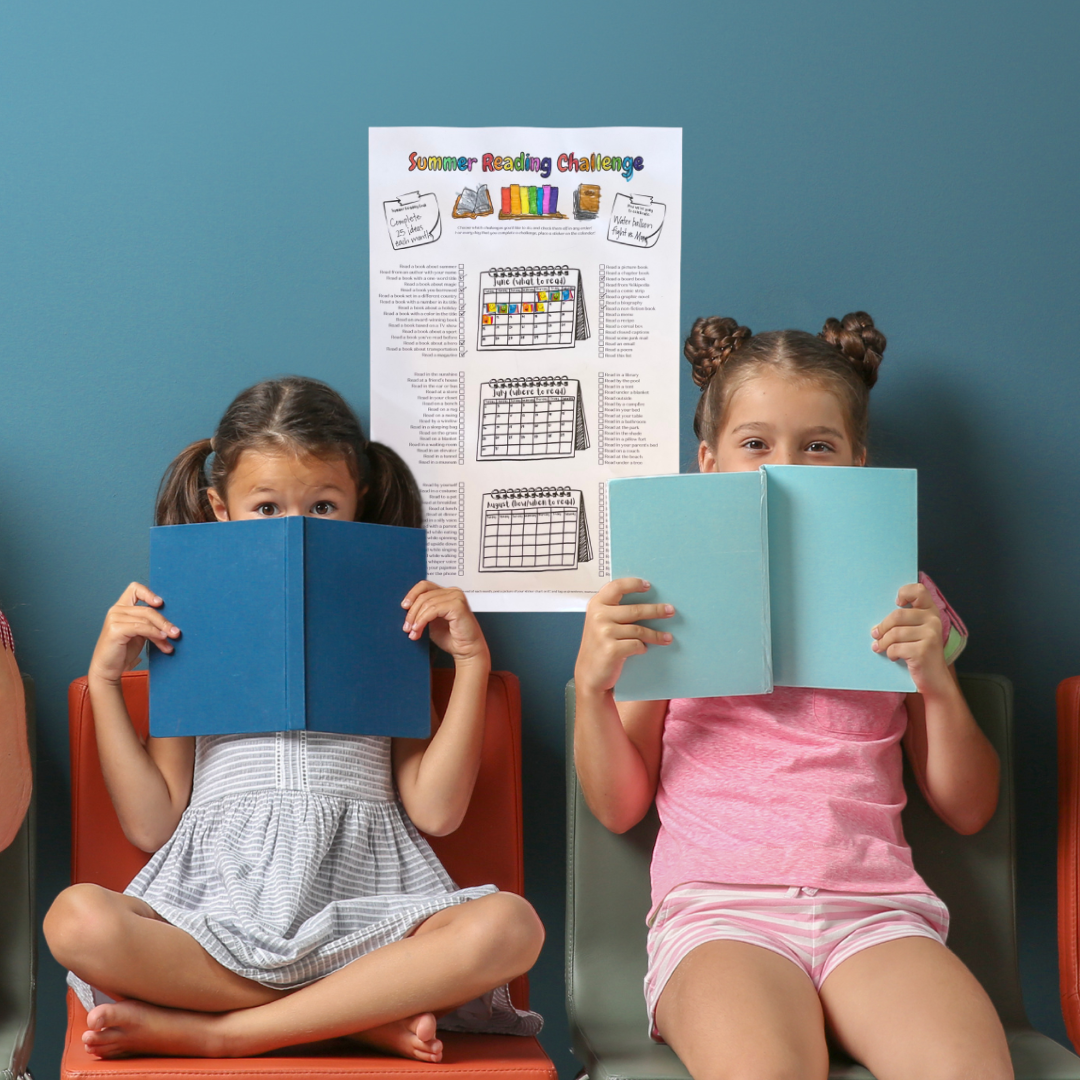 Two elementary school aged girls smile over the tops of their books as they sit near the Summer Reading Challenge calendar.