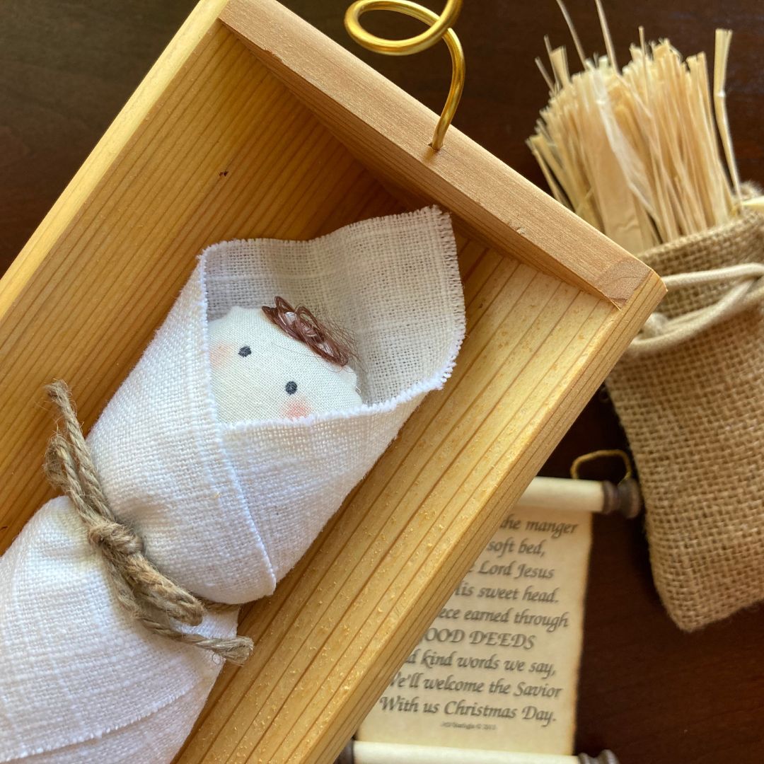 A cloth baby Jesus doll lies in a wooden manger, next to a burlap bag of straw and a scroll ornament that explains the Good Deeds Manger tradition.