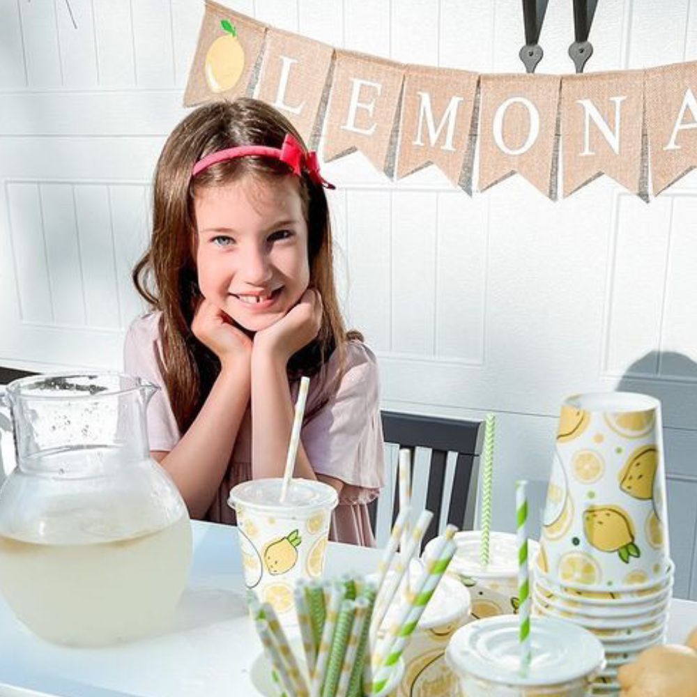 A close up photo of a girl sitting at a lemonade stand and smiling.
