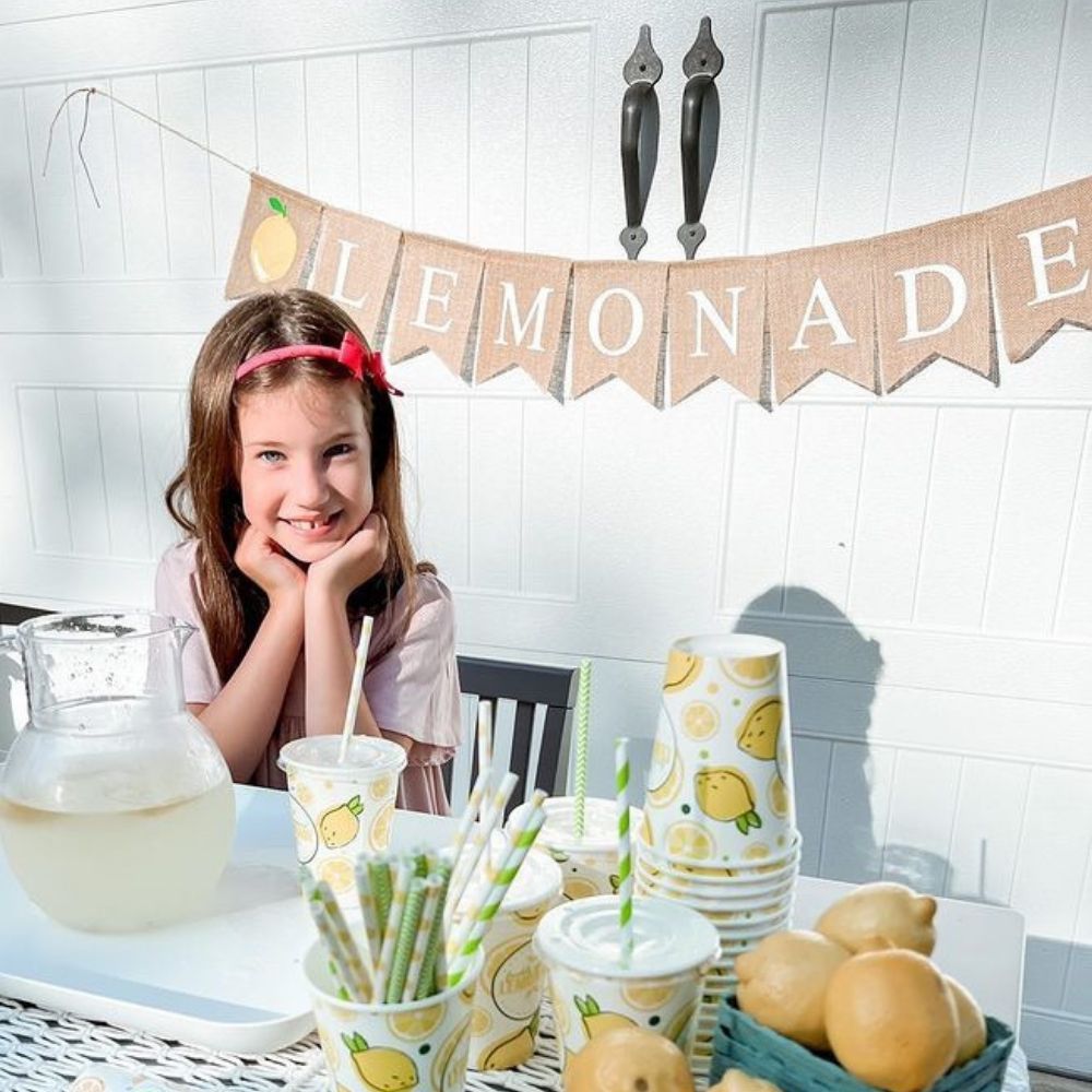 A girl sits at a lemonade stand, with a burlap pennant banner behind her that spells out Lemonade.