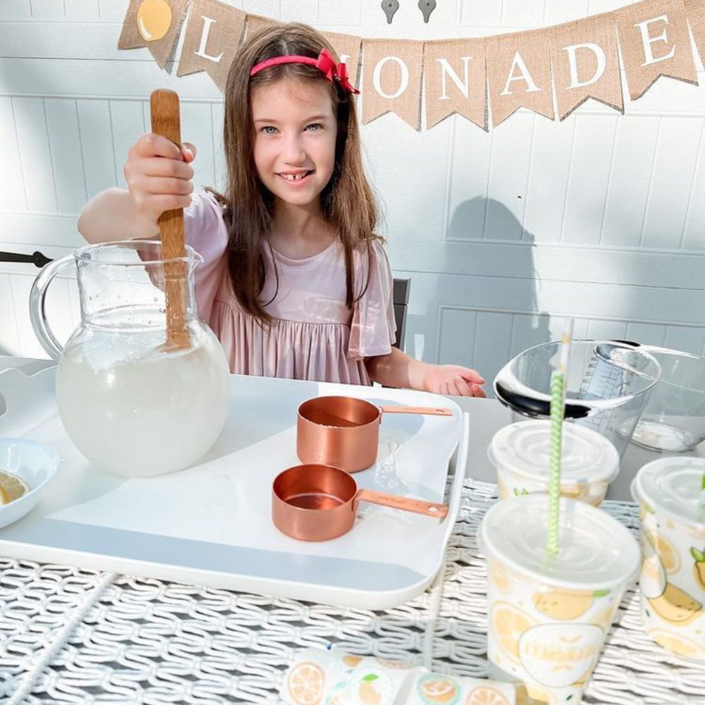 A girl mixes lemonade in a glass pitcher at a lemonade stand.