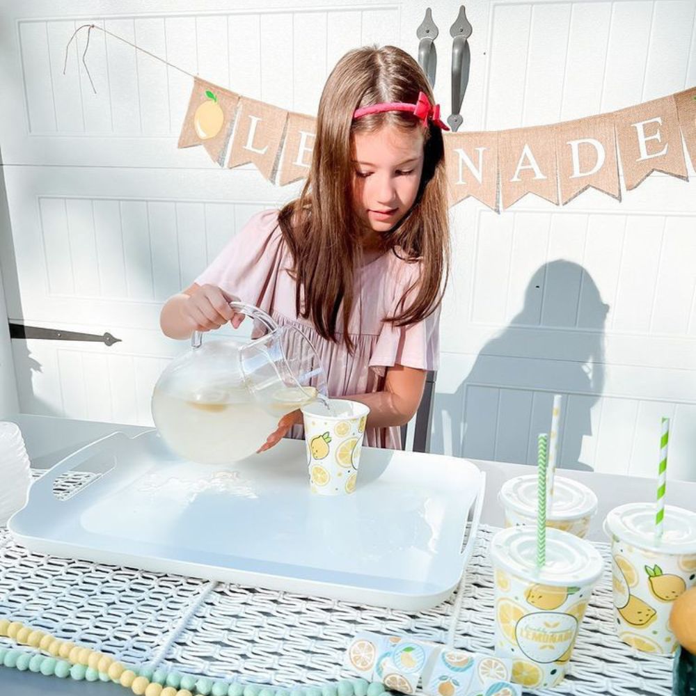 A girl pours lemonade into a paper cup with lemon/lemonade decorations on it at a lemonade stand.