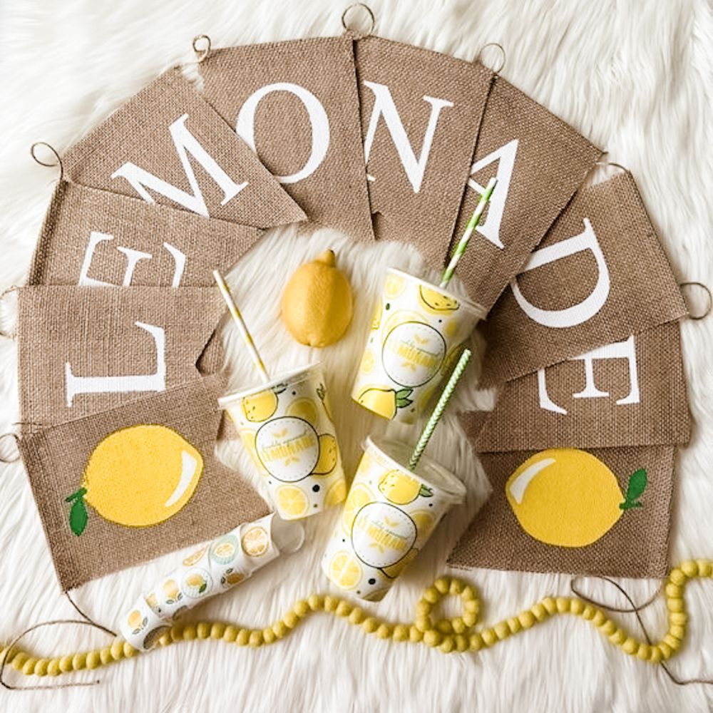 A burlap pennant banner spelling out Lemonade with a lemon at each end lays on a table surrounding some lemonade cups.